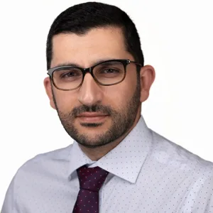 Professional headshot of a man with dark hair, beard and glasses wearing a white dotted shirt and burgundy tie