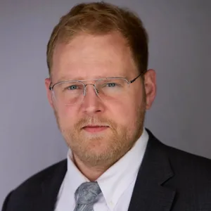 Professional headshot of a man with reddish-blonde hair, beard and glasses wearing a dark suit and gray tie