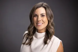 Professional headshot of a woman with shoulder-length brown wavy hair wearing a white high-neck top