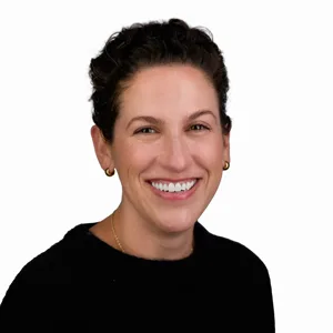 Professional headshot of a woman with short dark curly hair wearing a black top and gold earrings