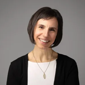 Professional headshot of a woman with short dark hair wearing a black cardigan over a white top with a leaf pendant necklace